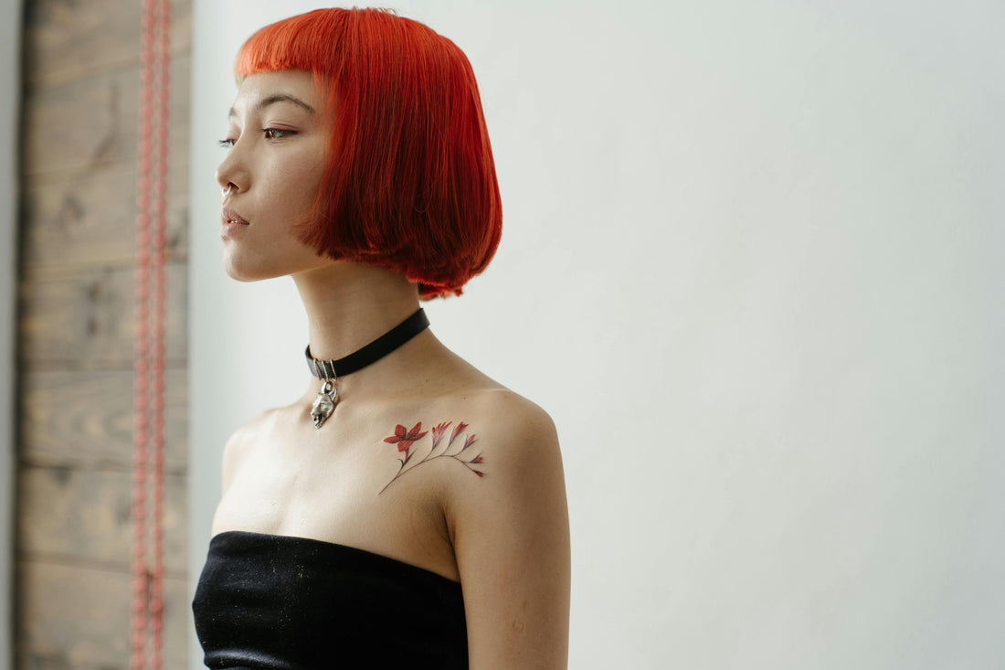 A woman with a bright red bob haircut is wearing a black choker and a strapless black top. She stands in profile against a white background, showcasing the vibrant tattoo ink of red flowers on her left shoulder.