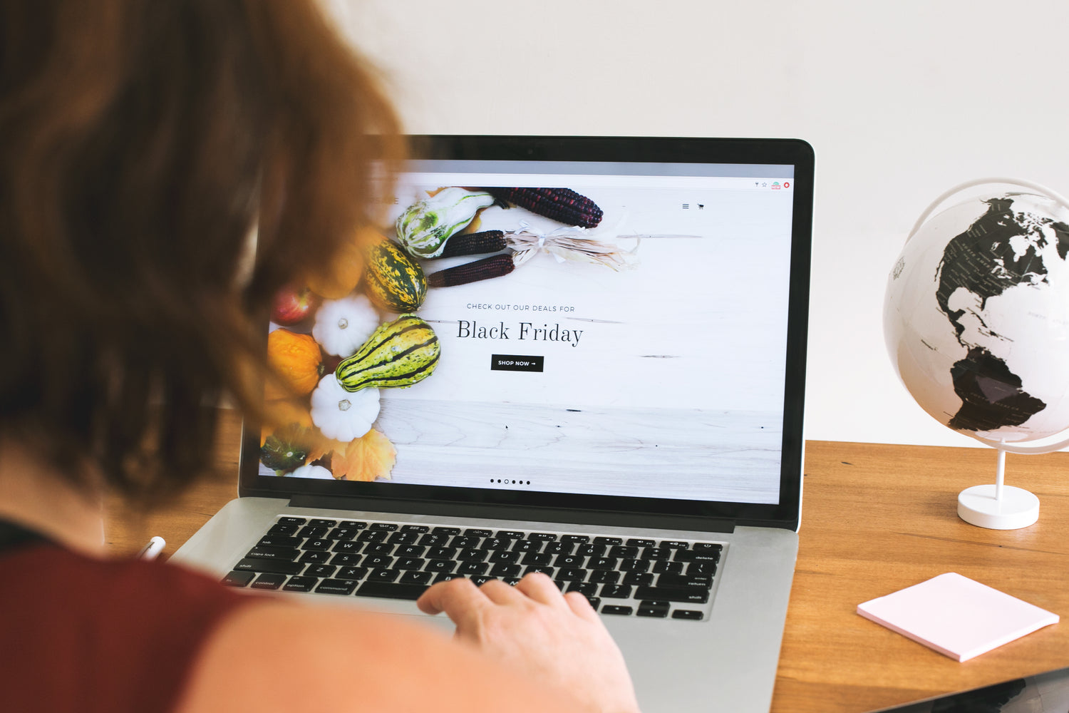  A person with shoulder-length hair, showcasing a hint of tattoo ink on their wrist, uses a laptop displaying a Black Friday sale advertisement. The ad features various colorful vegetables. A small globe and a sticky note are placed on the wooden table next to the laptop.