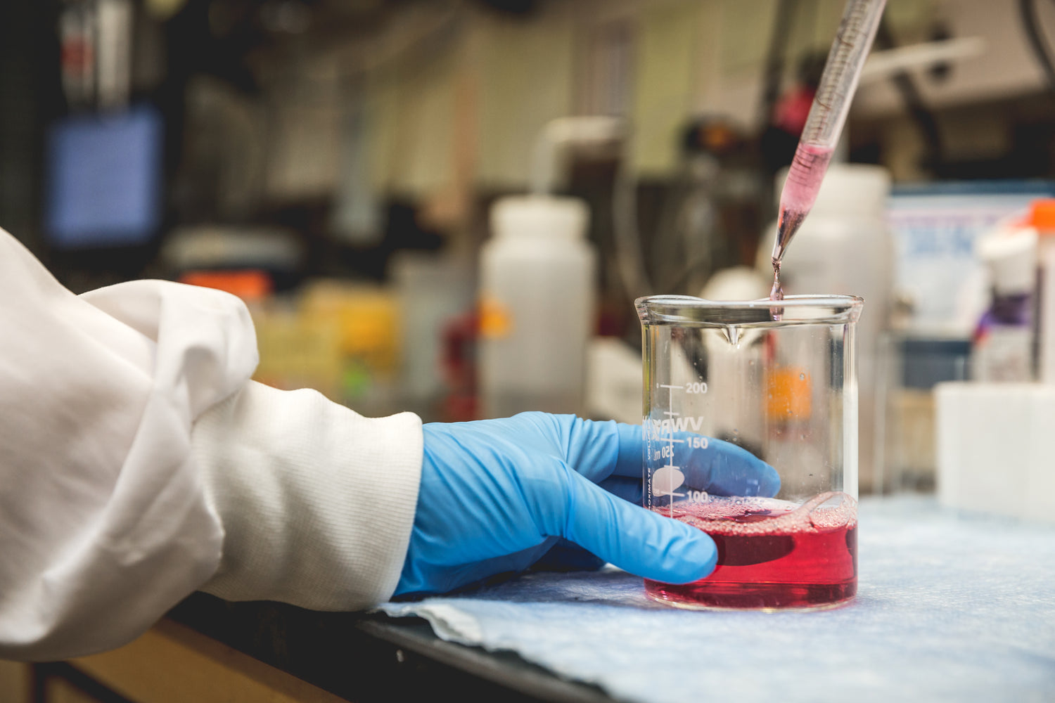 A person wearing a white lab coat and blue gloves uses a pipette to transfer a red liquid, resembling tattoo ink, into a beaker on a lab bench. The background shows various laboratory equipment and supplies.